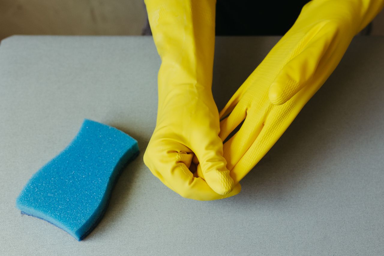services-03 Close-up of hands wearing yellow gloves next to blue sponge on gray surface.